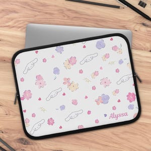 Puede incluir: Una funda para portátil blanca con un patrón repetitivo de caras de perro de dibujos animados con orejas largas, pequeños corazones rosas y flores estilo acuarela en tonos rosa, morado y amarillo. El nombre Alyssa aparece en rosa abajo.