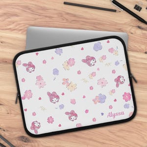 Puede incluir: Una funda para portátil blanca con un patrón repetitivo de cabezas de conejo de dibujos animados rosas, varias flores estilo acuarela en tonos rosa, amarillo y morado, pequeños corazones rojos, las letras HK y el nombre Alyssa en la esquina.