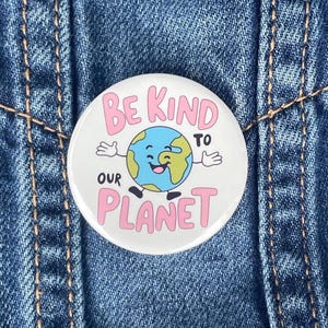 May include: White button with a pink and blue design. The button features a cartoon earth with a smiling face and the text "Be Kind to Our Planet".