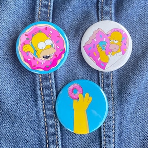 May include: Three round pinback buttons featuring the cartoon character Homer Simpson. The first button shows Homer swimming in a pink donut with sprinkles. The second button shows Homer eating a donut with a pink glaze. The third button shows Homer's hand holding a pink donut.