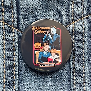 May include: A black button featuring a colourful image of a horror movie character holding a knife, a woman and a child reading a book, and a pumpkin. The text on the button reads "Halloween Safety A Sitter's Guide".