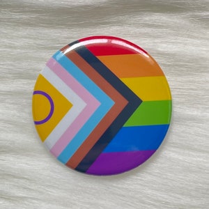 May include: A round button with a rainbow pride flag design. The flag includes the colors red, orange, yellow, green, blue, indigo, and violet. The flag also includes a black and brown stripe, a light blue stripe, and a pink stripe. The flag is surrounded by a white circle.