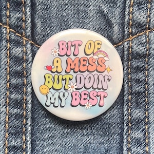 May include: A round button with the text "BIT OF A MESS BUT DOIN MY BEST" in colorful, retro-style lettering. The button features a rainbow, flowers, a heart, and a smiley face on a pastel tie-dye background.