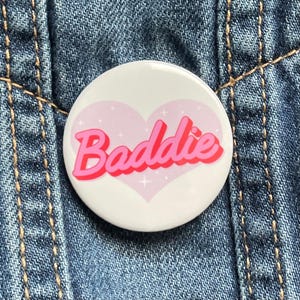May include: A white button with a pink heart and the word "Baddie" in pink and red lettering. The button is set against a blue denim background.