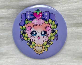 May include: A circular pinback button featuring a light purple background with a central illustration of a hamster. The hamster is framed by a green leaf wreath, a yellow sunflower at the bottom, and a purple bow with a pink strawberry at the top.