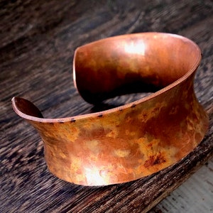 May include: A wide copper cuff bracelet with a hammered finish. The bracelet has a warm, reddish-brown colour and a slightly irregular shape.