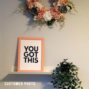 May include: A framed print with the text "YOU GOT THIS" in navy blue letters, set against a white background, and framed in a peach-colored frame. Above, a floral wreath with pink, white, and gray flowers. A small plant is on the right.