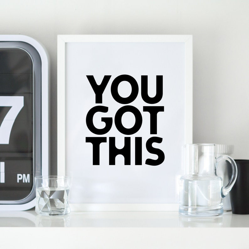May include: A white framed print with the text "YOU GOT THIS" in bold black letters. The print is displayed on a white shelf, alongside a geometric glass, a pitcher, a black cup, and a digital clock.