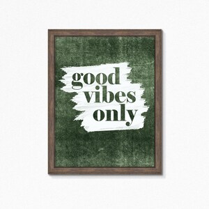 May include: Framed wall art featuring the phrase "good vibes only" in black lettering. The text is set against a white brushstroke on a textured, dark green backdrop. The frame is a dark brown colour.