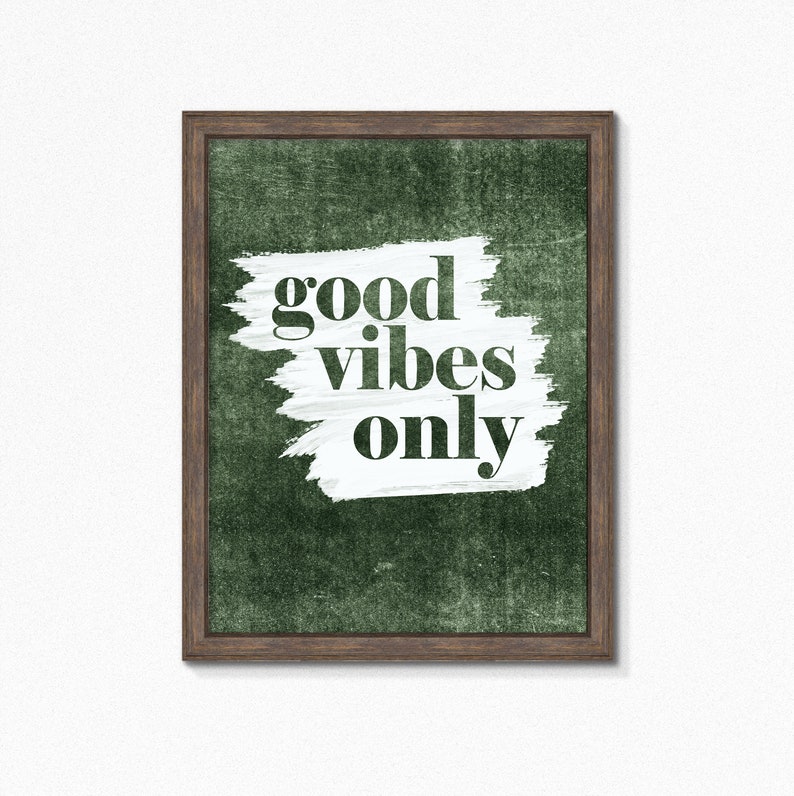 May include: Framed wall art featuring the phrase "good vibes only" in black lettering. The text is set against a white brushstroke on a textured, dark green backdrop. The frame is a dark brown colour.