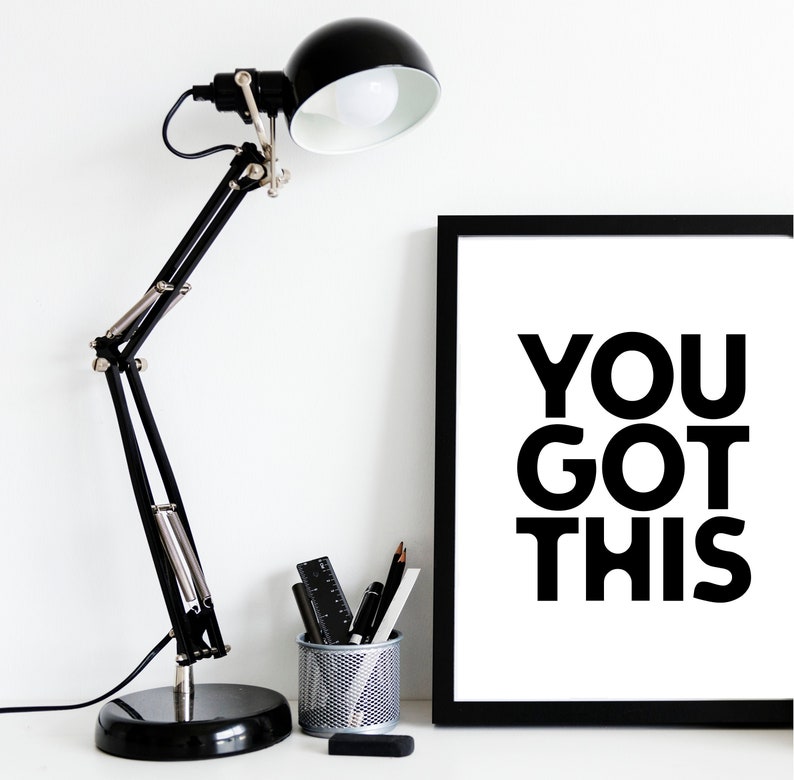 May include: A black desk lamp with an adjustable arm and a round base is positioned next to a framed print. The print has the words "YOU GOT THIS" in bold, black letters. A small metal pencil holder with pens, pencils, and a ruler is also visible.