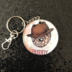 May include: A round, clear keychain with a cartoon image of Freddy Krueger wearing a brown fedora. The keychain has a silver metal ring and clasp.