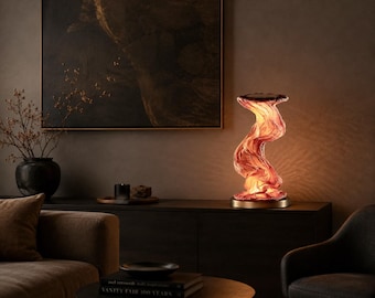 Puede incluir: Una lámpara de mesa con un cuerpo de vidrio translúcido de tonos rojo anaranjado con remolinos y una forma fluida y orgánica. La lámpara está montada sobre una base metálica circular y emite un brillo cálido que resalta la superficie texturizada.