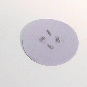 May include: A pale lilac, circular object featuring four silver staples arranged in a cross formation. The object is set against a white background.