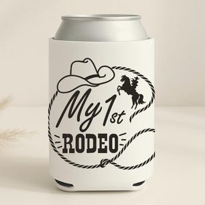 May include: White can cooler with a silver top, featuring a black cowboy hat, a horse and rider silhouette, and the text "My 1st Rodeo" in a black font. The design is encircled by a black rope.