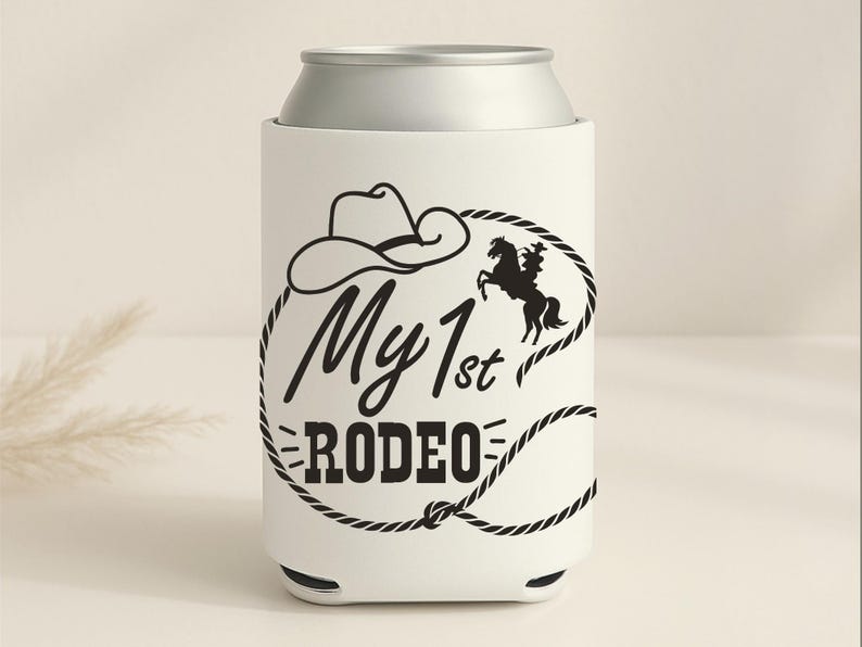 May include: White can cooler with a silver top, featuring a black cowboy hat, a horse and rider silhouette, and the text "My 1st Rodeo" in a black font. The design is encircled by a black rope.