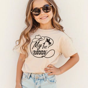 May include: A beige t-shirt with the words "My 1st Rodeo" in black, with a cowboy hat, horse, and lasso design. The shirt is worn by a young person wearing sunglasses and light blue jeans.