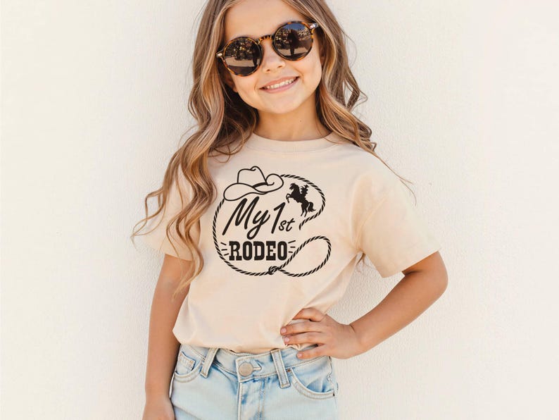 May include: A beige t-shirt with the words "My 1st Rodeo" in black, with a cowboy hat, horse, and lasso design. The shirt is worn by a young person wearing sunglasses and light blue jeans.