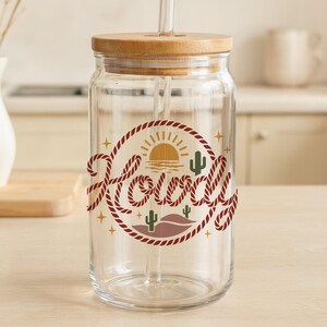 May include: A clear glass drinking jar with a wooden lid and straw. The jar features a circular design with the word "Howdy" in a rope-like font, along with a desert landscape illustration in shades of brown, green, and yellow.