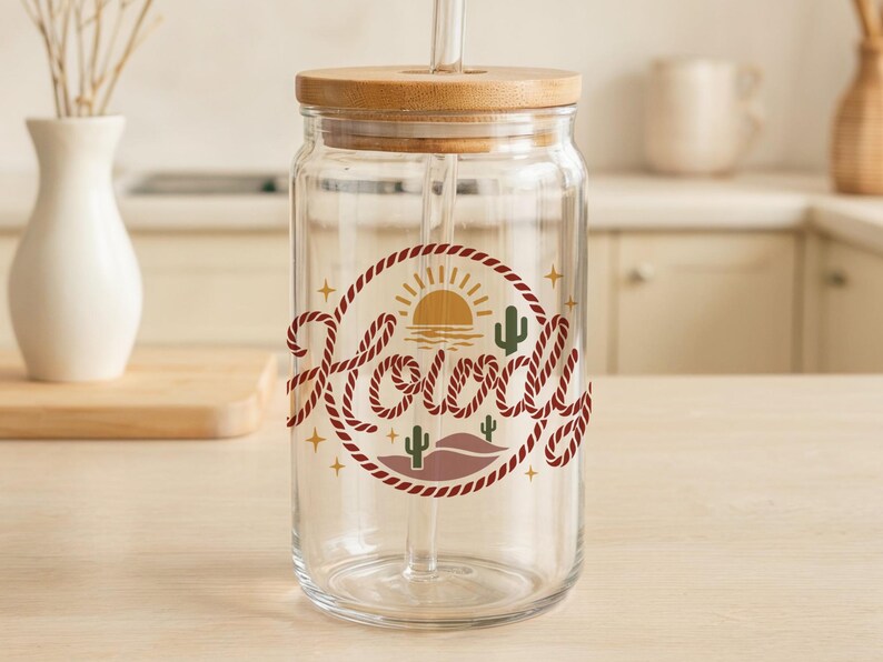 May include: A clear glass drinking jar with a wooden lid and straw. The jar features a circular design with the word "Howdy" in a rope-like font, along with a desert landscape illustration in shades of brown, green, and yellow.