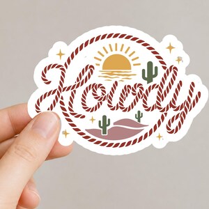 May include: A white sticker with the word "Howdy" in a rope-style font, encircled by a rope border. The design includes a yellow sun, cacti, and stars, evoking a Western theme.