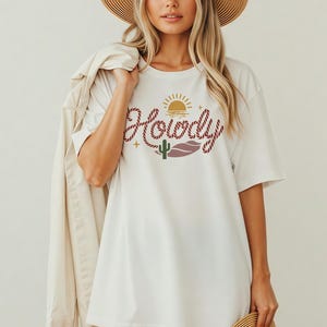 May include: White t-shirt with a graphic design featuring the word "Howdy" in a rope-like font, a sun, a cactus, and a lip shape. The shirt is a casual, oversized style.