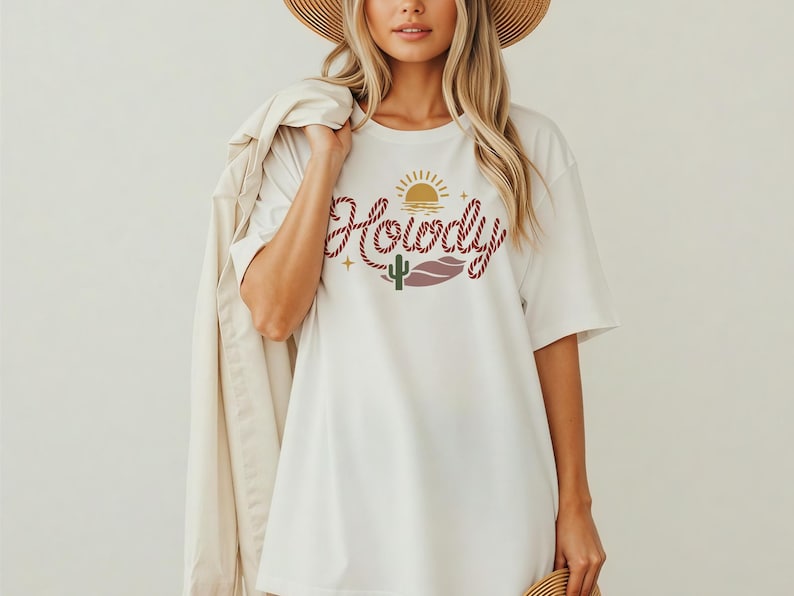 May include: White t-shirt with a graphic design featuring the word "Howdy" in a rope-like font, a sun, a cactus, and a lip shape. The shirt is a casual, oversized style.
