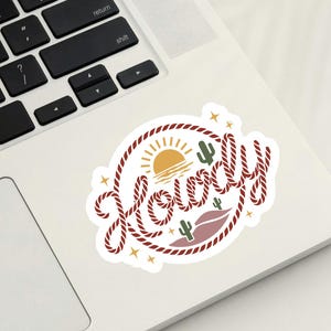 May include: A white sticker with a western-themed design. The sticker features the word "Howdy" in a rope-style font, surrounded by a rope border. The design includes a sun, cacti, and stars.