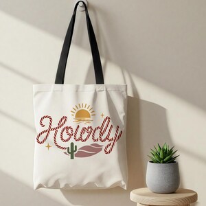 May include: A white tote bag with black handles, featuring the word "Howdy" in a rope-like font. The design includes a sun, cactus, and desert landscape elements. A small potted plant sits nearby.