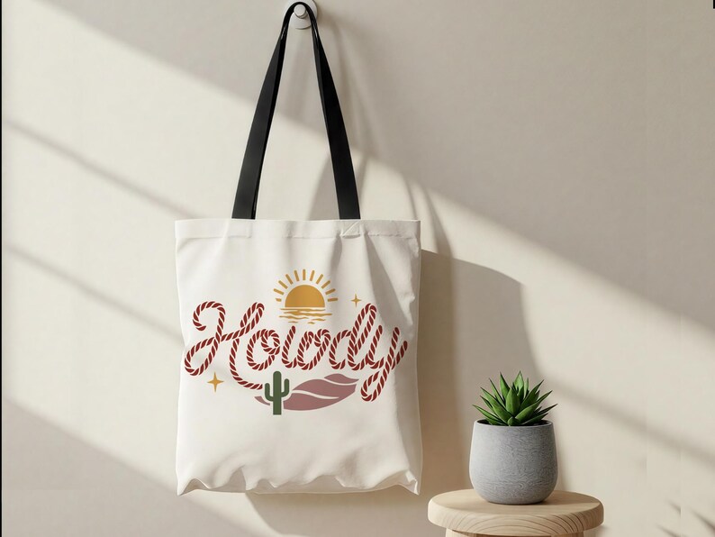 May include: A white tote bag with black handles, featuring the word "Howdy" in a rope-like font. The design includes a sun, cactus, and desert landscape elements. A small potted plant sits nearby.