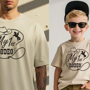 May include: Two beige t-shirts, one worn by an adult and the other by a child. Both shirts have a black graphic with a cowboy hat, horse, and the text "My 1st Rodeo". The shirts are short-sleeved.