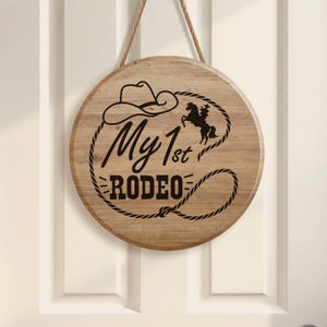 May include: A circular wooden sign with the words "My 1st Rodeo" in black script, a cowboy hat, and a horse silhouette. The sign is suspended by a rope and is mounted on a white door.