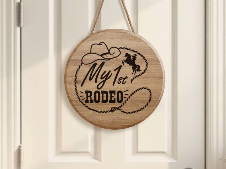 May include: A circular wooden sign with the words "My 1st Rodeo" in black script, a cowboy hat, and a horse silhouette. The sign is suspended by a rope and is mounted on a white door.