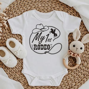May include: White baby bodysuit featuring the text "My 1st Rodeo" in black, with a cowboy hat and horse illustration. Includes white crochet baby shoes and a crochet bunny rattle.