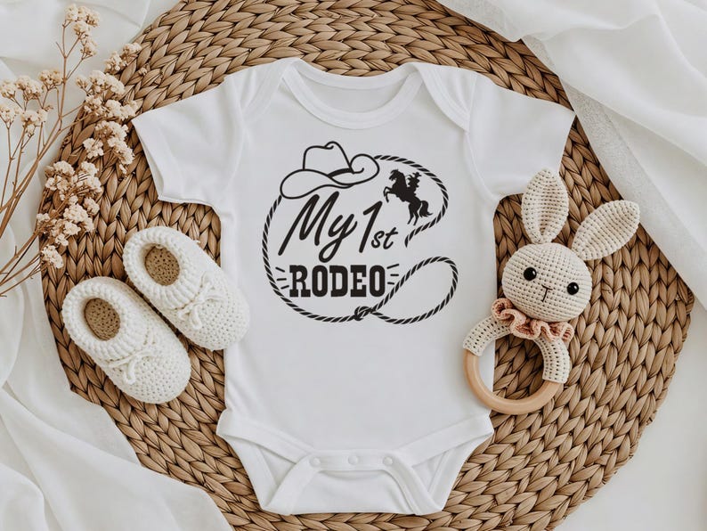 May include: White baby bodysuit featuring the text "My 1st Rodeo" in black, with a cowboy hat and horse illustration. Includes white crochet baby shoes and a crochet bunny rattle.