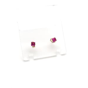 May include: A pair of small stud earrings. Each earring features a square, bright pink gemstone set in a gold-colored metal. The earrings are displayed on a clear plastic card against a white background.