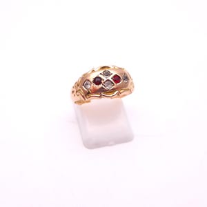 May include: A gold ring with a domed shape, featuring a pattern of alternating red and clear gemstones. The ring has an ornate, engraved design along the band and edges, set against a white background.