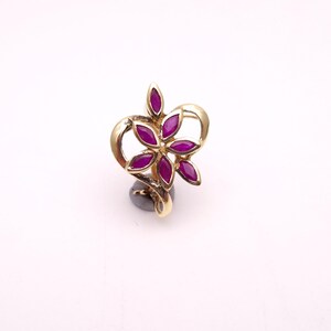 May include: A gold-coloured ring featuring a flower design with six deep magenta petals. The petals are arranged around a central point, and the ring has a heart-shaped design. The ring is set on a dark grey base.