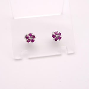 May include: A pair of silver stud earrings. Each earring has a flower design with six deep pink gemstones around a central, smaller, clear gemstone. The earrings are displayed on a white background.