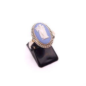 May include: Silver ring with an oval blue cameo depicting a classical figure in white relief. The ring has a detailed beaded border and a plain band. The ring is displayed on a black stand against a white background.