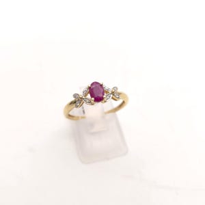 May include: A gold ring with an oval ruby gemstone at its centre. The ring is embellished with small, round diamonds and has a floral pattern on either side of the ruby. It is displayed on a clear stand against a white backdrop.