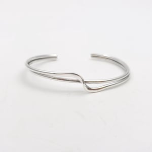 May include: A silver-toned metal cuff bracelet featuring a double-band design that curves and overlaps at the centre. The bracelet has an open back and a smooth, polished finish, creating a continuous, flowing shape across the wrist area.