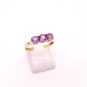 May include: A gold ring featuring three round, vibrant purple gemstones set in a row. The ring is displayed on a white stand against a plain white background, highlighting the jewelry's design and color.
