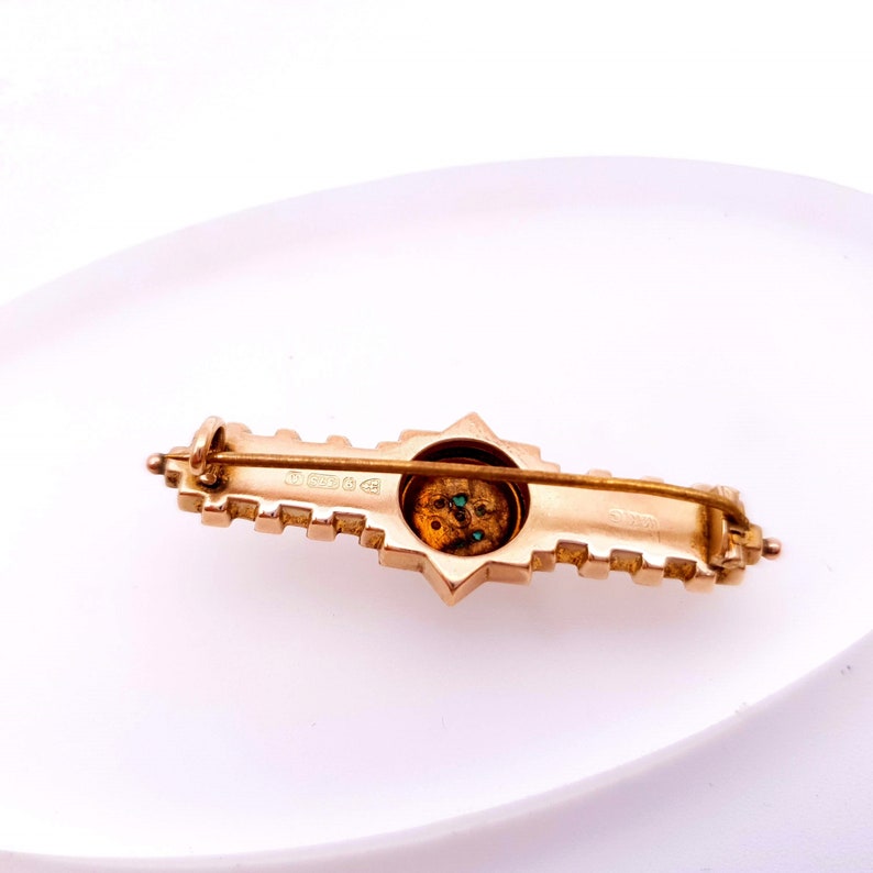 Pu&ograve; includere: Un fermaglio dorato con un design a forma di stella e un centro circolare con pietre verdi. Il retro del fermaglio ha un marchio.