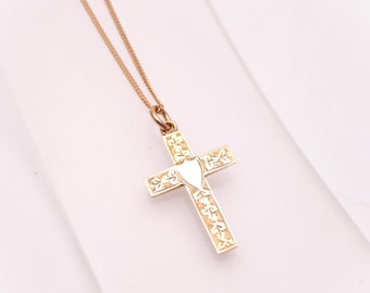May include: A gold-coloured cross pendant necklace. The cross has a shield-shaped centre and floral detailing on the arms. The chain is delicate and gold-toned, creating a classic and elegant look.