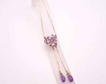 May include: A gold-toned necklace featuring a floral cluster of purple gemstones. The necklace has a delicate chain and two teardrop-shaped purple gemstones dangling at the end. The jewelry is displayed on a white stand.