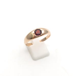 May include: A polished gold ring featuring a single, round, faceted dark red garnet gemstone set in a star-shaped bezel. The band is smooth and tapers slightly towards the back, showcasing a classic design with a warm, metallic lustre.