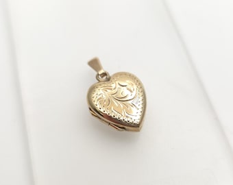 May include: A gold-colored heart-shaped locket pendant with an ornate floral design. The locket has a small loop at the top for a chain. The pendant is approximately 1 inch in length and width.
