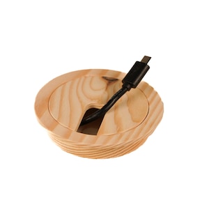 Puede incluir: Una tapa redonda de gestión de cables de madera con un cable negro que la atraviesa.