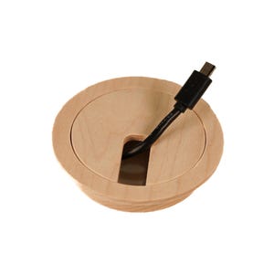 Puede incluir: Una tapa redonda de gestión de cables de madera con un cable negro que la atraviesa. La tapa tiene un acabado liso de color marrón claro y un borde ligeramente elevado.